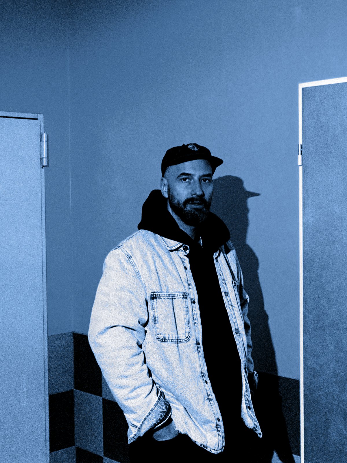 Mohig in a cap, a black hoodie and a light denim jacket, standing between two doors